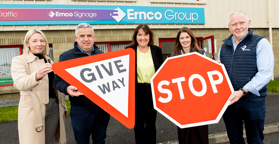 Three women and two men are standing in front of Ernco Group's office and holding road signs