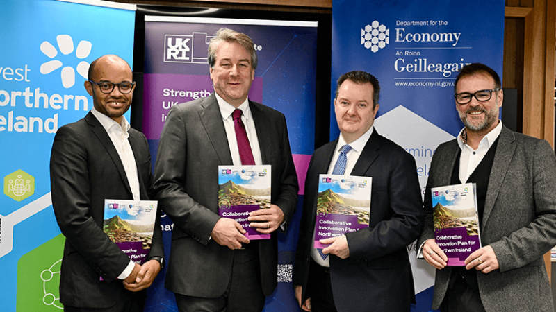 Four men standing in front of company pop-up branding, each holding a copy of the Collaborative Innovation plan for Northern Ireland